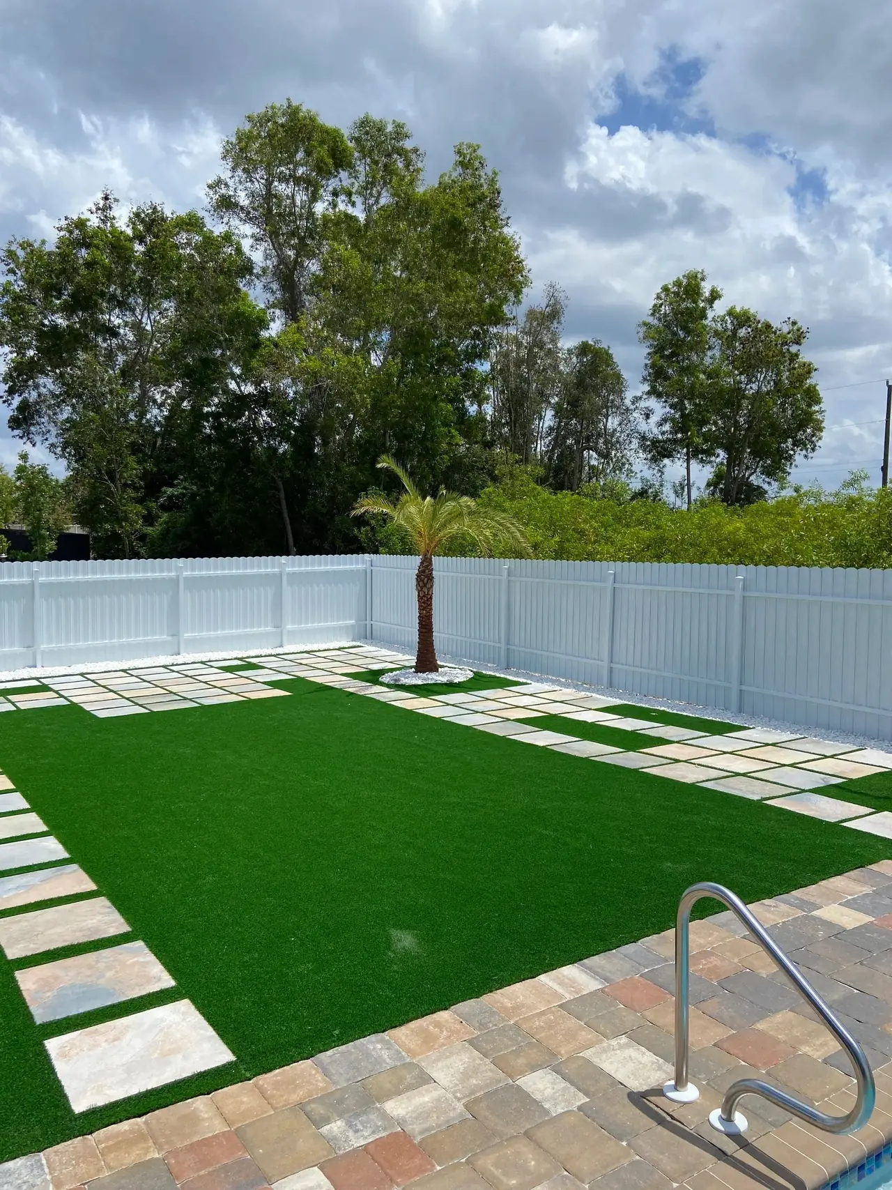 Patio de grama sintética con losas de piedra y una palmera, junto a una piscina con borde de adoquines